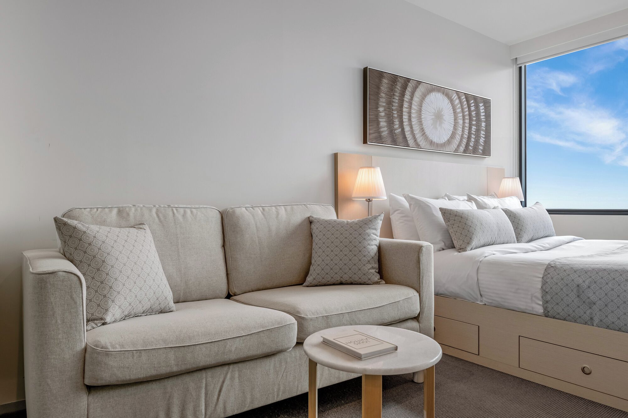 A modern hotel room with a sofa, a bedside table with a book, a bed with a patterned bedspread, a wall-mounted lamp, and a large abstract painting.