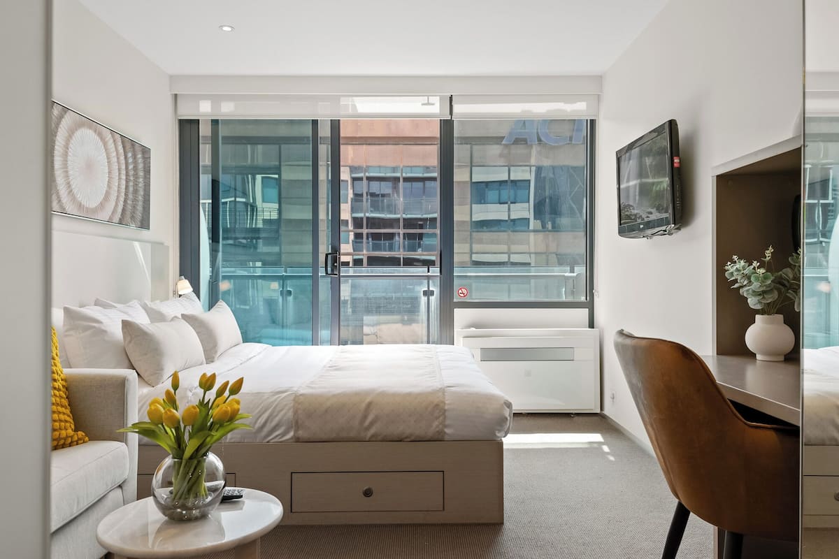 Ett modernt hotellrum med en säng, ett skrivbord, en stol, en TV och ett stort fönster med utsikt över staden.