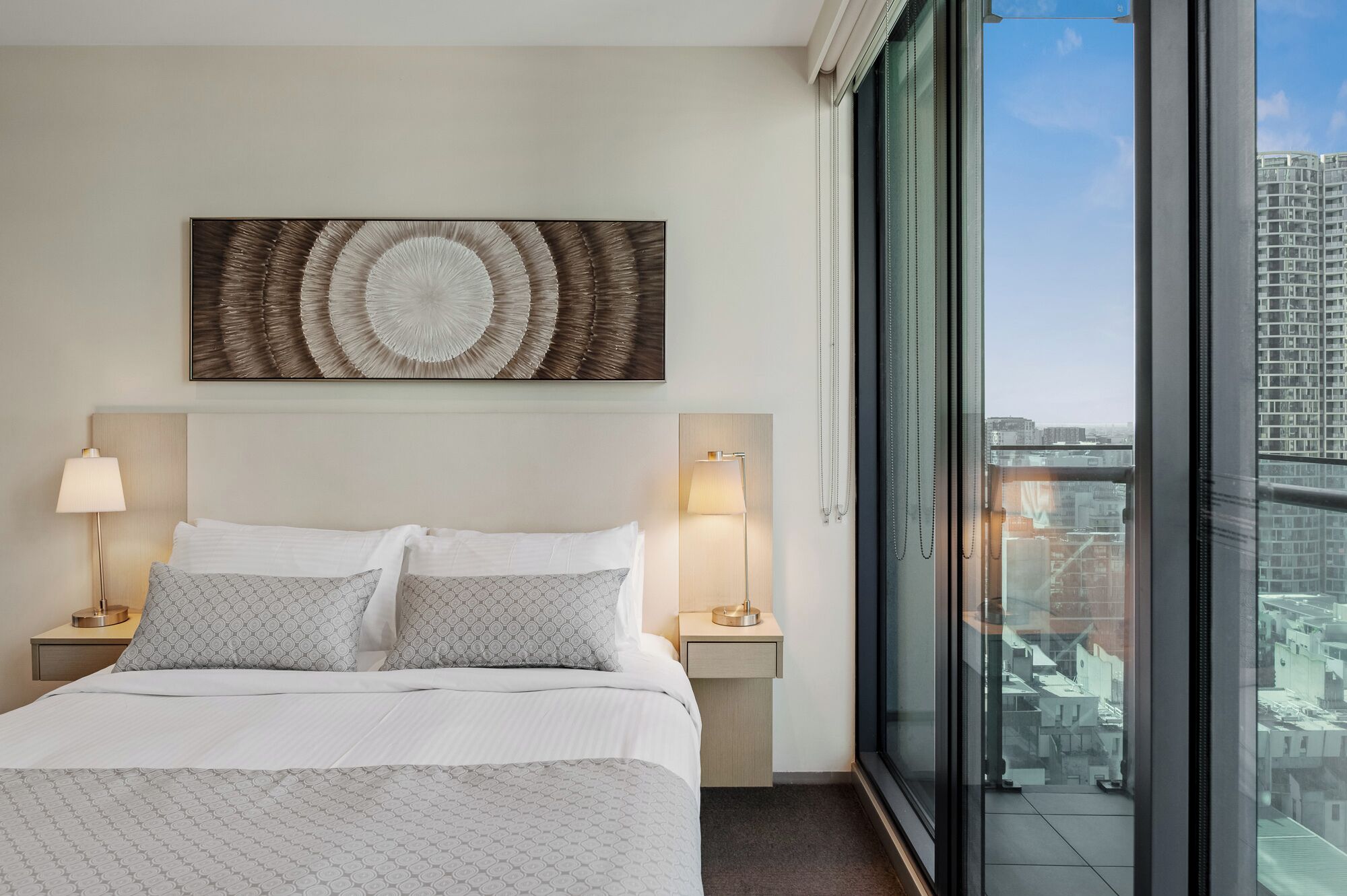 Ein Hotelzimmer mit einem großen Bett, zwei Nachttischlampen, einem Wandbild und Blick auf die Stadt durch ein großes Fenster.