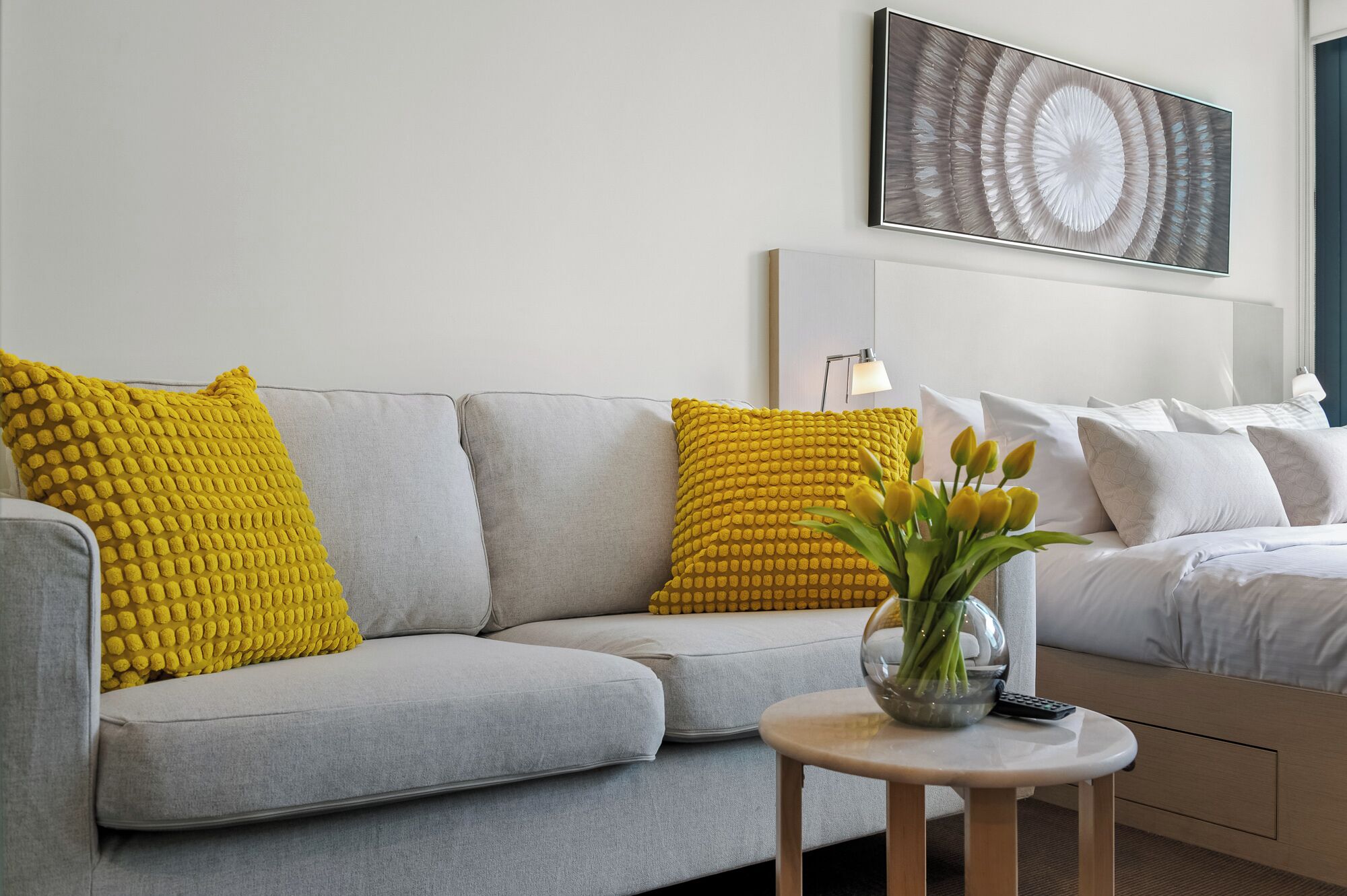 Una sala de estar moderna con un sofá gris, almohadones amarillos con textura, una mesita redonda con un florero y una cama con ropa de cama blanca.