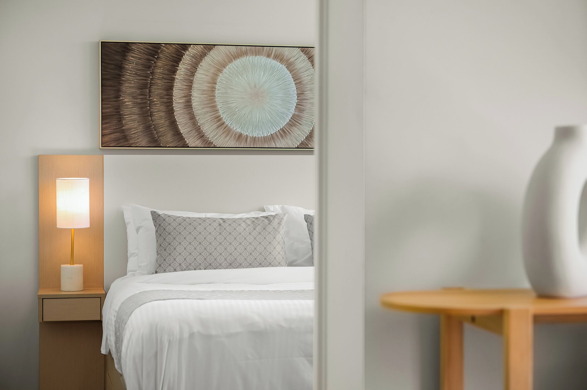 Et pænt indrettet hotelværelse med seng, natlampe, et billedværk på væggen og et lille bord med en vase.