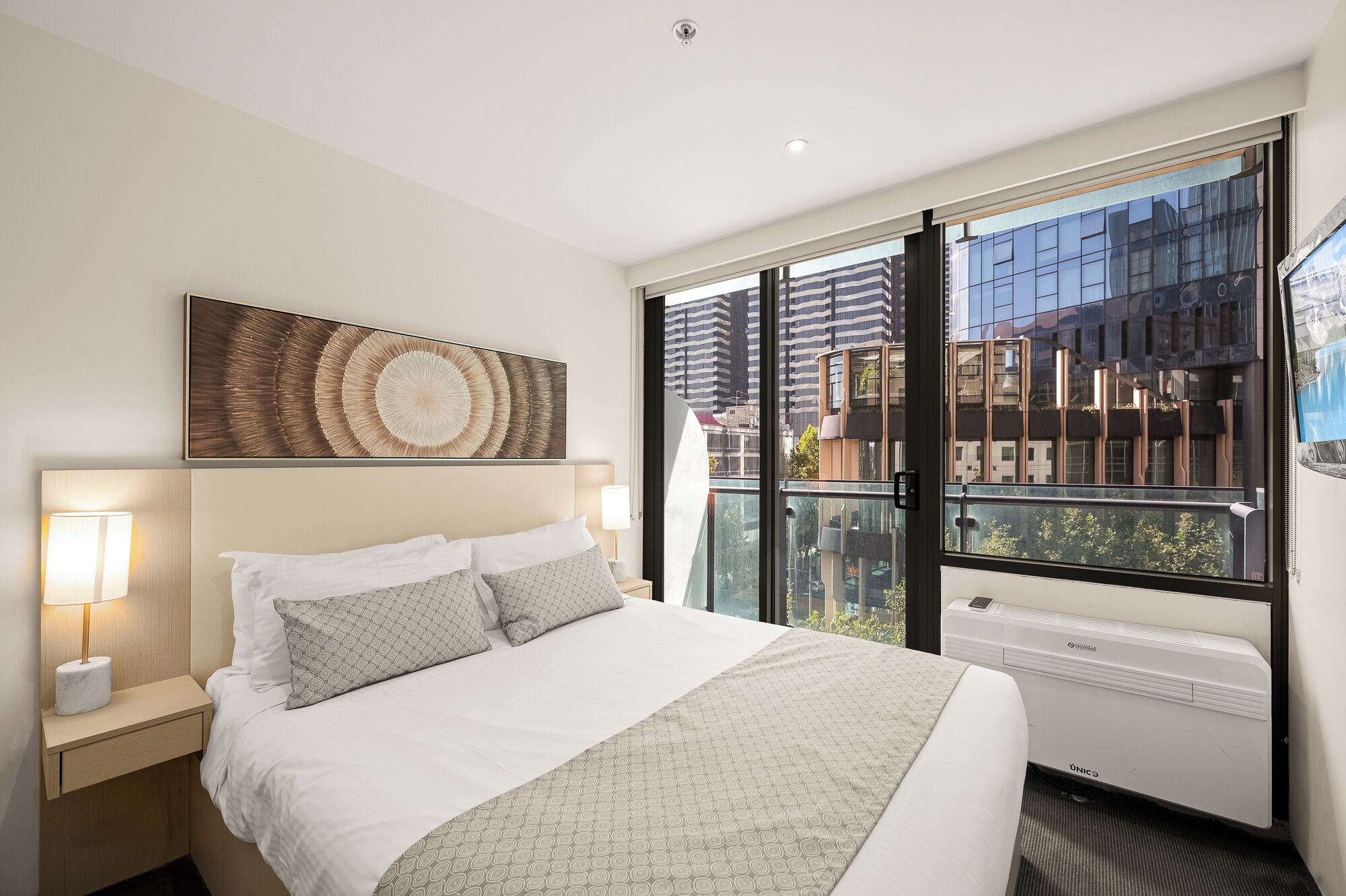 A hotel room with a bed, a wall-mounted artwork, a window with a view of buildings, and a radiator.