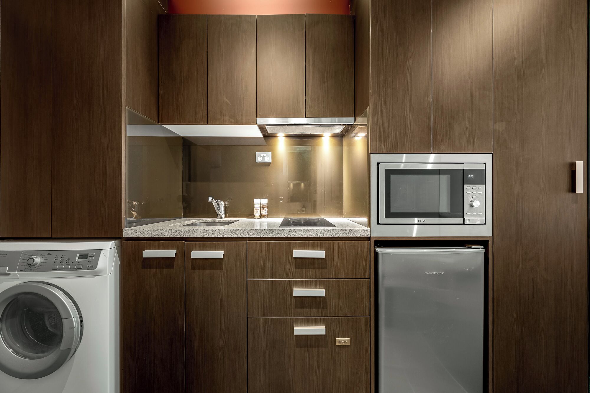 A compact kitchen with wooden cabinets, a stainless steel microwave, a built-in washing machine, and a sink.