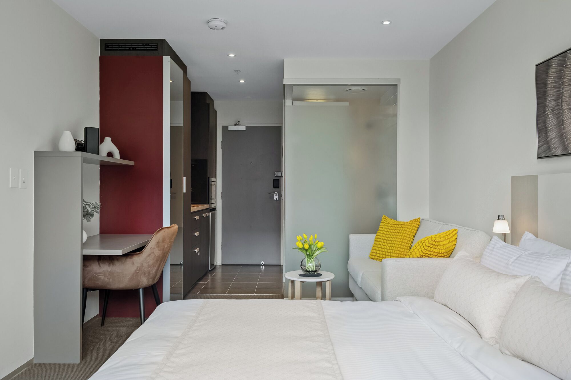 Ein modernes Hotelzimmer mit einem großen Bett, einem Schreibtisch mit Stuhl, einem kleinen runden Tisch mit einer Vase und einer Tür zu einem Badezimmer.