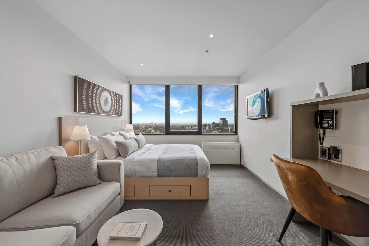 Ett modernt hotellrum med en säng, en soffa, ett soffbord, ett skrivbord och en stol. Det finns ett stort fönster med utsikt över staden, en platt-TV och en telefon på skrivbordet.