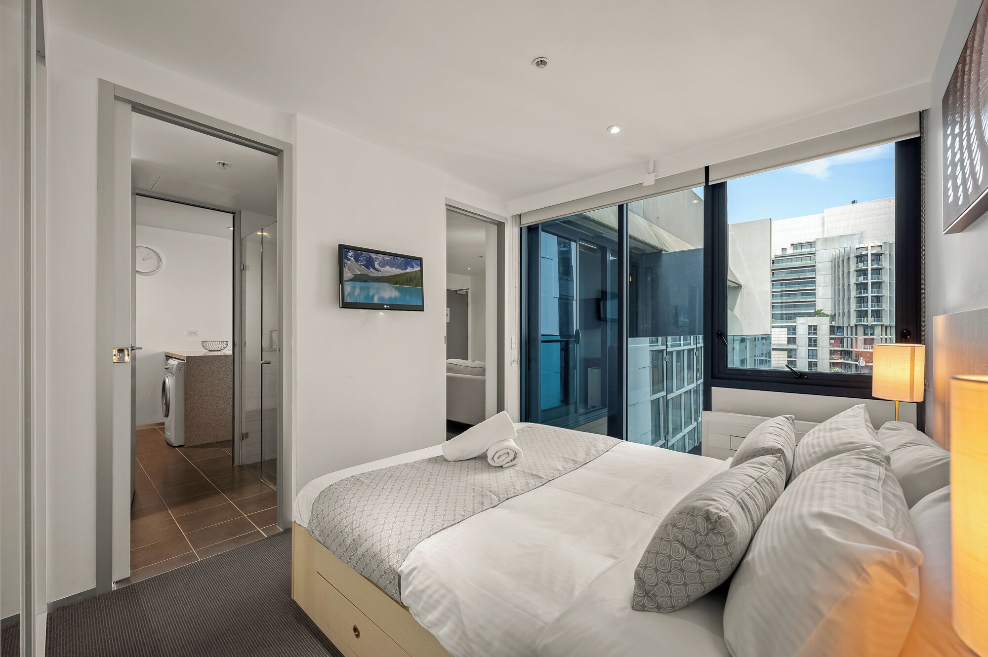 Une chambre d’hôtel moderne avec un grand lit, une vue sur la ville par une porte vitrée et une petite salle de bain équipée d’un lave-linge.