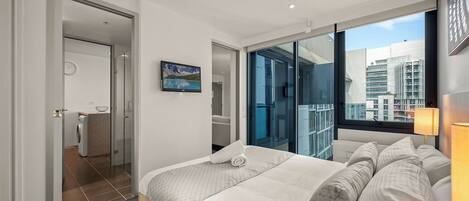 A modern hotel room with a large bed, a view of the city through a glass door, and a small bathroom with a washing machine.