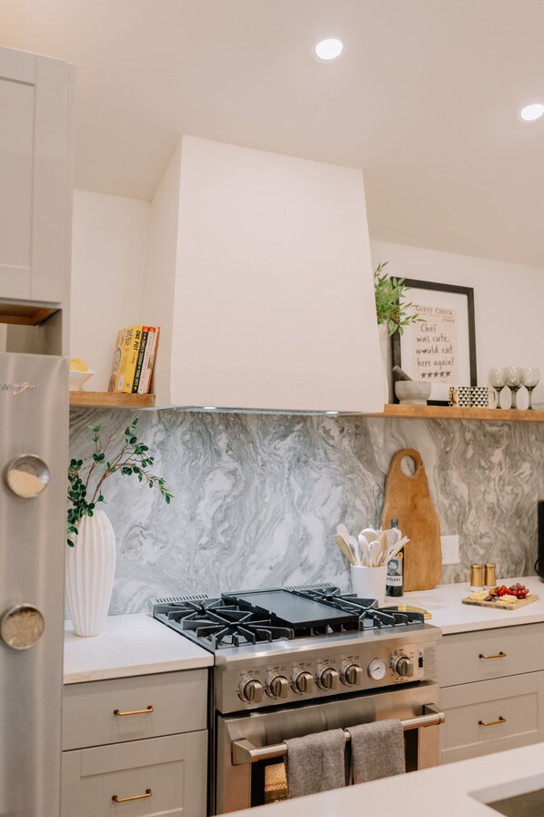 A modern kitchen with a built-in stove, oven, and white marble backsplash. There are wooden shelves above the counter holding books and decorative items.