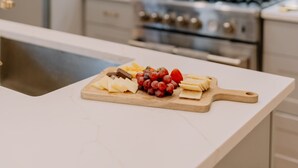 Una cocina con salpicadero de mármol, una tabla de madera con queso y uvas, y una estufa de acero inoxidable.