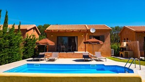 A pool area with lounge chairs and umbrellas, surrounded by buildings with terracotta roofs. - Rhea Villa in Kefallonia (Kefalonia)