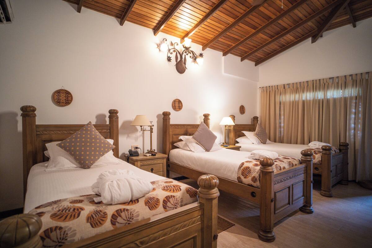 A hotel room with two wooden beds, each with a headboard, bedside tables, lamps, and a large window with curtains.