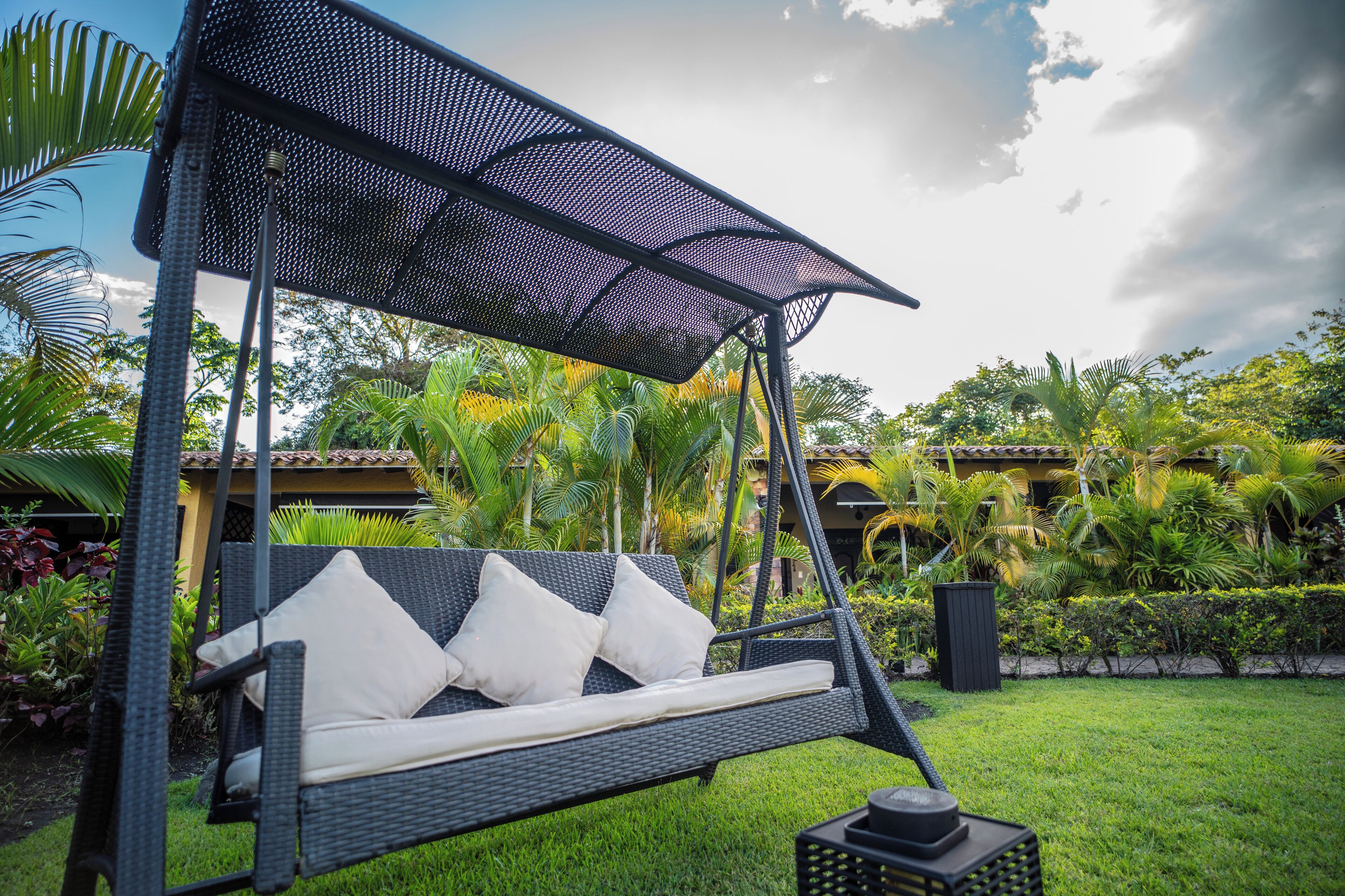 A black wicker swing chair with white cushions and a canopy, set in a lush garden with tropical plants and a building in the background.