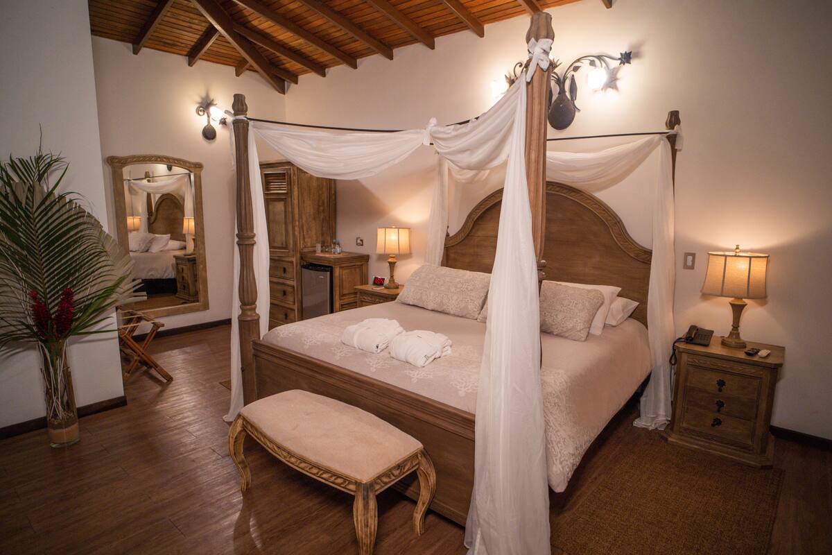 A four-poster canopy bed with a wooden headboard, bedside tables with lamps, a bench, and a large mirror.