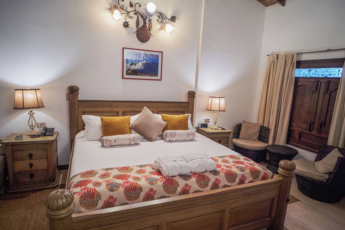 A neatly made bed with a floral bedspread, two bedside tables with lamps, a wooden headboard, a wall-mounted picture, and wicker chairs.