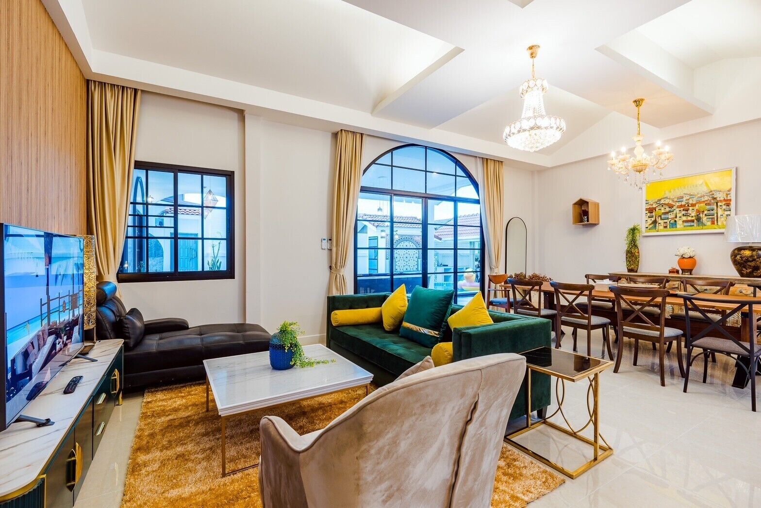 A modern living room with a sofa, coffee table, and TV. There are large windows and a chandelier.