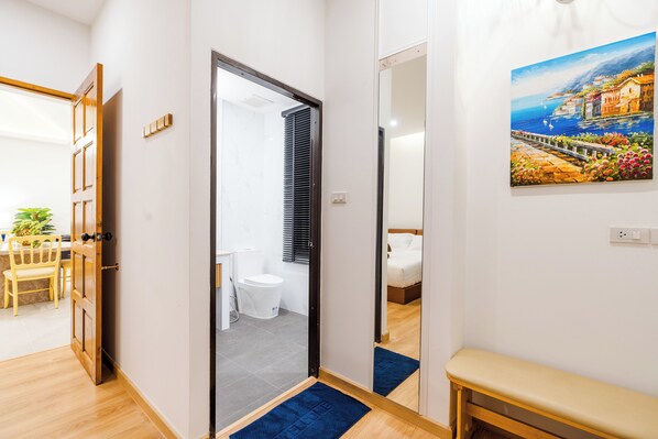 A modern hotel room with a wooden floor, a bathroom with a toilet and sink, a bed, and a painting of a coastal scene.