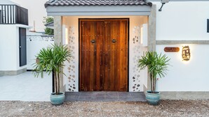 A traditional wooden door with a metal handle, flanked by two potted plants, leading to a building with a tiled roof and a signboard.