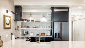 A modern kitchen with a white countertop, black cabinetry, and a stainless steel refrigerator. There are shelves above the counter holding various kitchenware and a coffee machine.