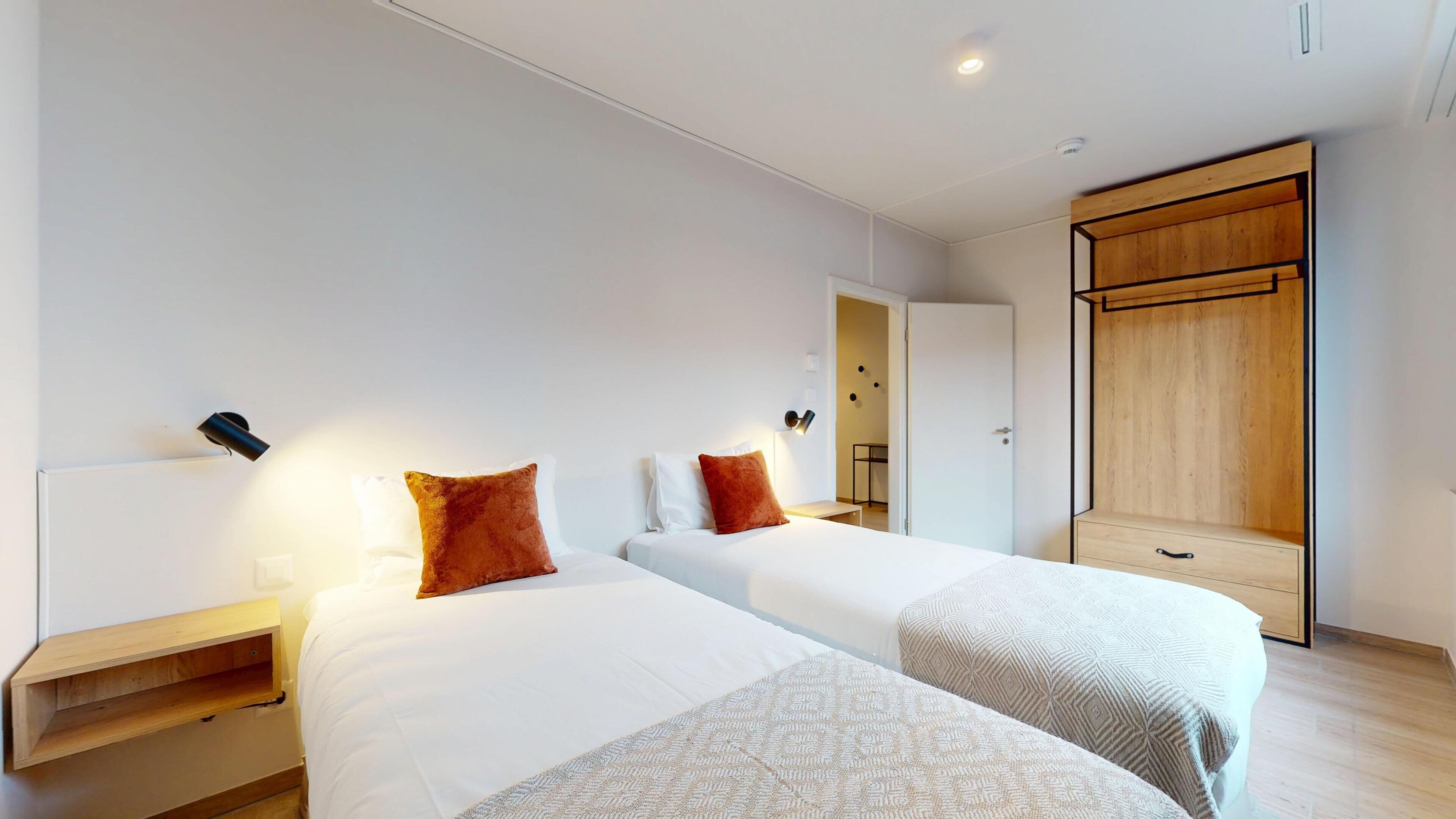 Quarto de hotel moderno com duas camas, um guarda-roupa de madeira e uma pequena mesa de cabeceira.