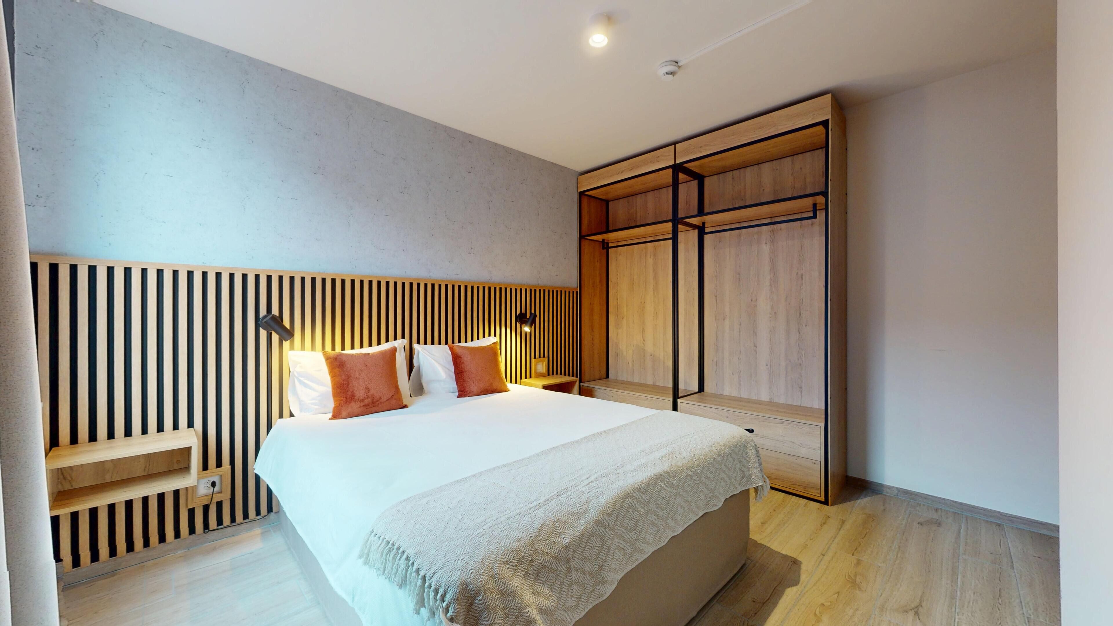 A modern bedroom with a wooden slatted headboard, a bed with white linens and two orange pillows, a bedside table with a lamp, and a wooden wardrobe.