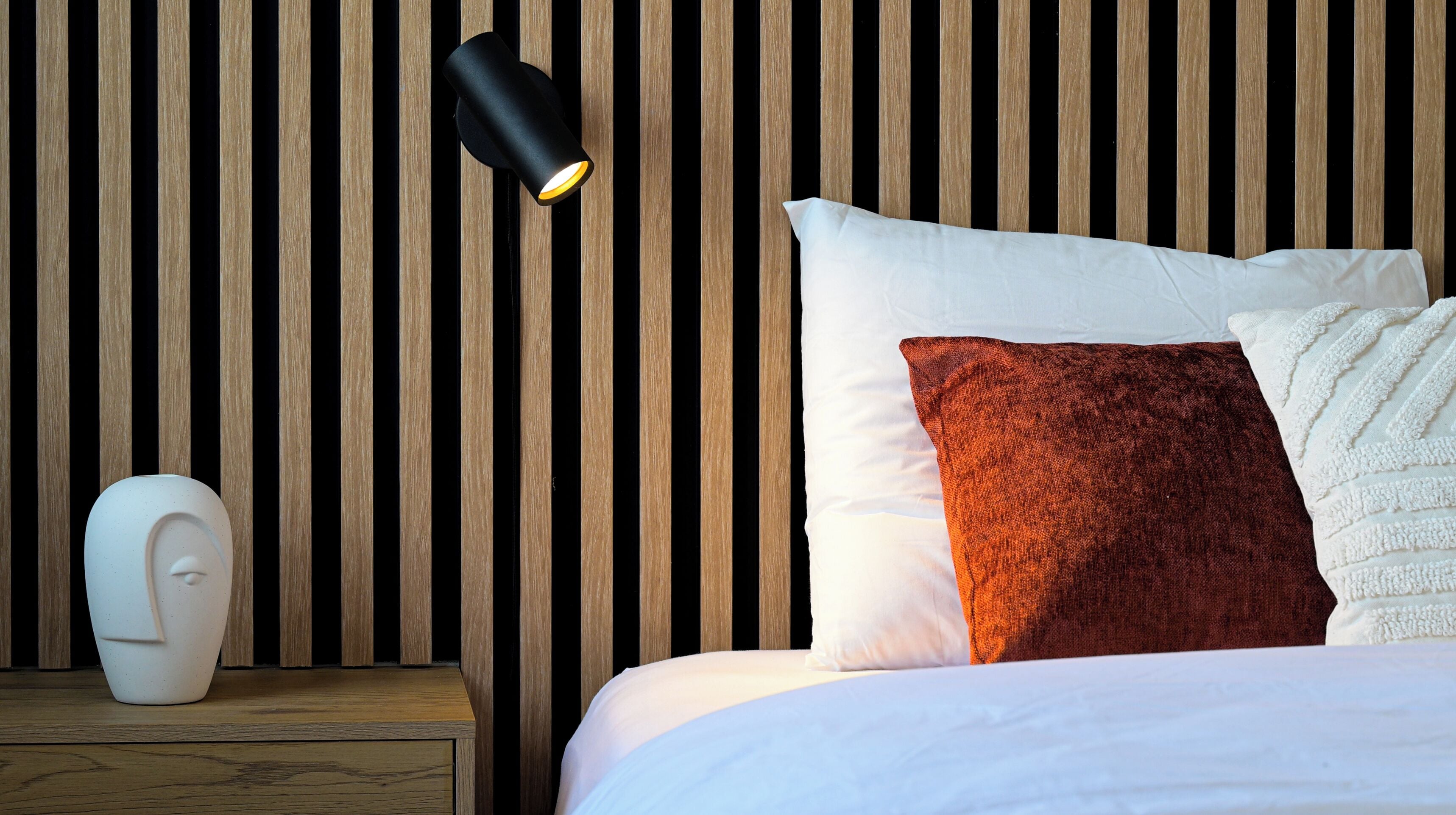 Un lit bien fait, avec des oreillers blancs et bruns, un vase blanc posé sur une table de chevet en bois et une lampe noire fixée au mur.