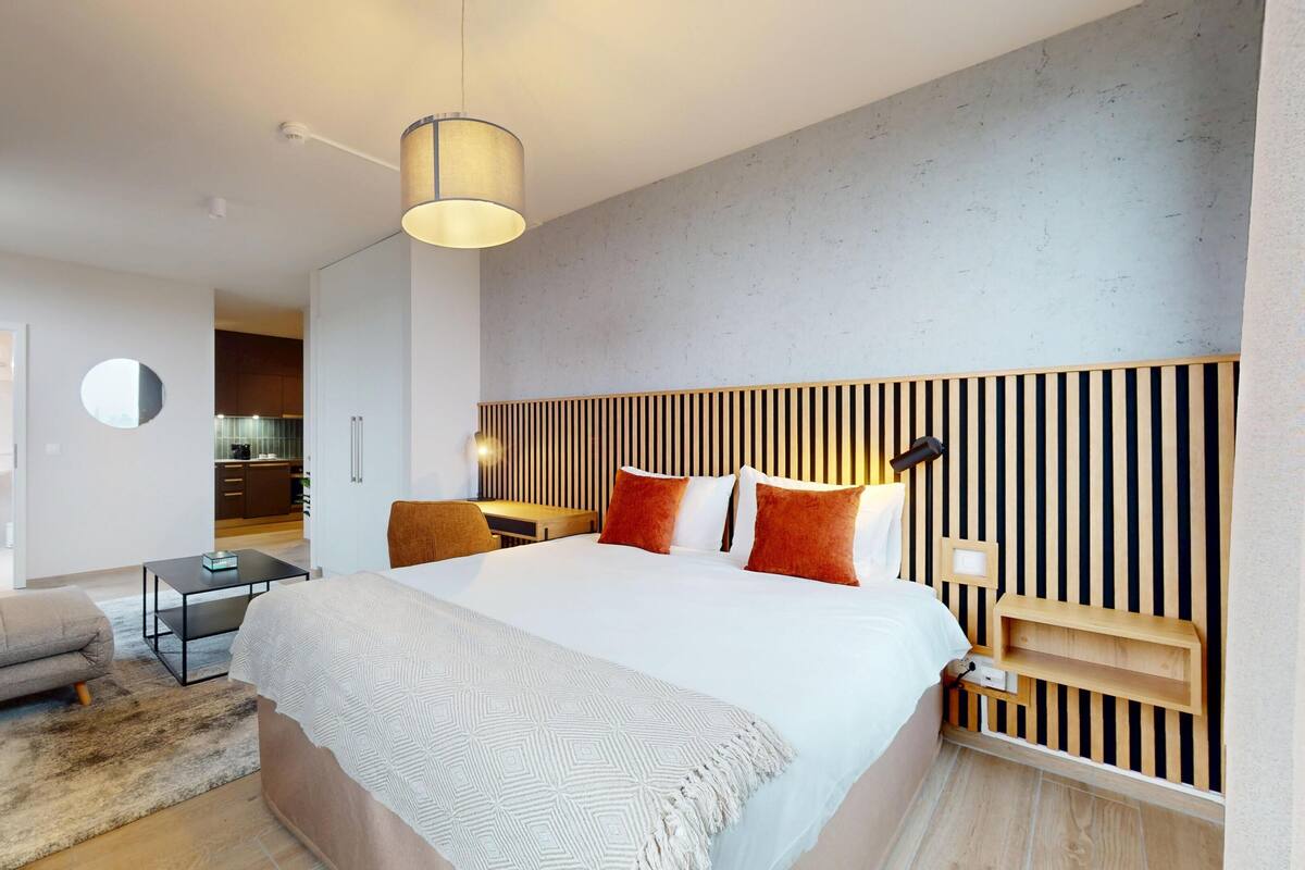 Habitación de hotel moderna con una cama grande, un escritorio, una silla y una pequeña kitchenette al fondo.
