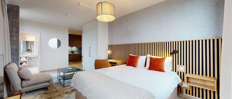 Habitación de hotel moderna con una cama grande, un escritorio, una silla y una pequeña kitchenette al fondo.