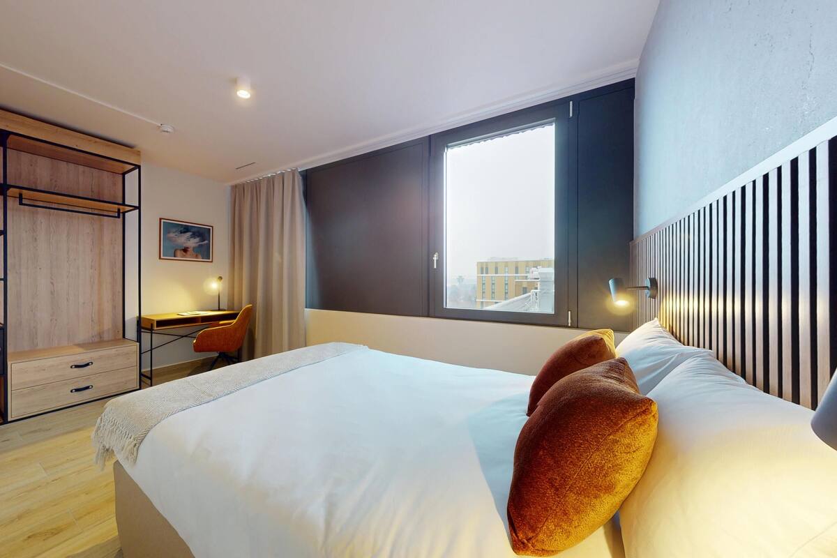 Una habitación de hotel con cama, escritorio, armario y ventana con vistas a la ciudad.