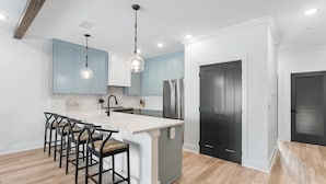 A modern kitchen with light blue cabinets, a white island, black chairs, stainless steel appliances, and a dark door. - Gus' Getaway (Port St. Joe)