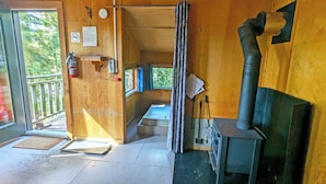 L’intérieur d’un chalet en bois, avec une cheminée, une fenêtre donnant sur les arbres, une porte avec un paillasson et un extincteur.