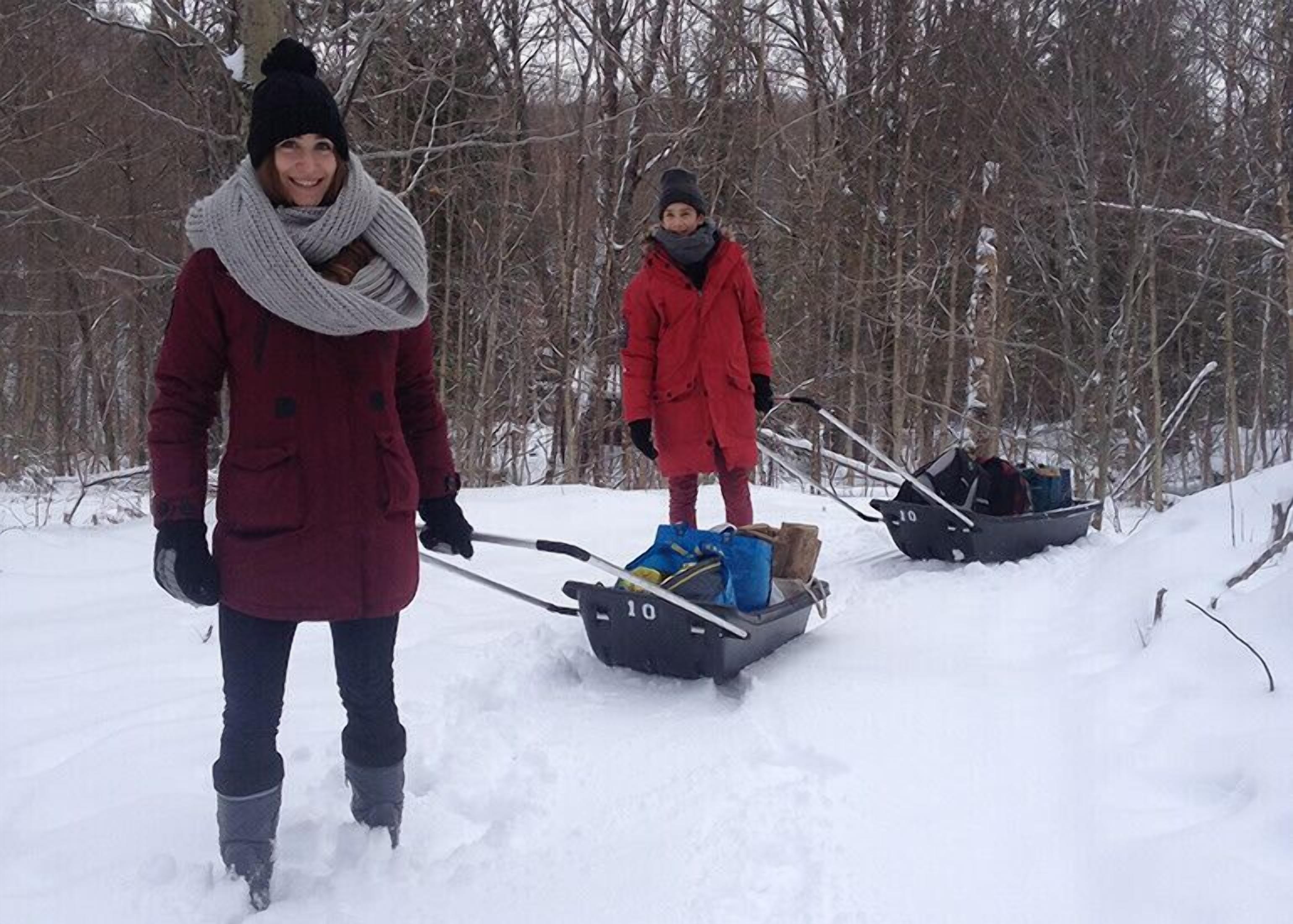 Two people in winter clothing pulling sleds through a snowy forest.
