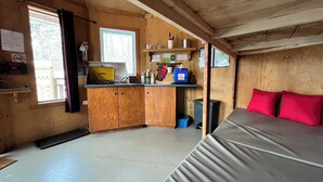 A compact cabin with a kitchenette, a bed with red pillows, and a window with a view of trees.