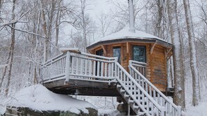 A wooden cabin with a staircase leading to a deck, surrounded by snow-covered trees.