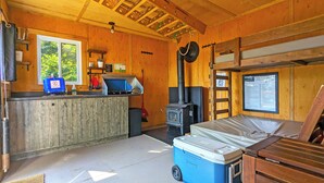 Interior de cabana com cozinha, lareira a lenha, beliche e janela com vista para o verde.