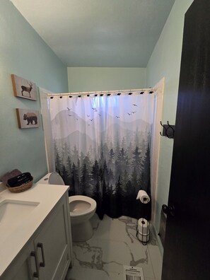 A bathroom with a shower curtain depicting a forest scene, a toilet, a sink with a basket of towels, and a wall-mounted towel rack. - River access, Quiet, Family and pet friendly. Best mountain views in the valley (Payson)