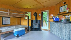 Interior de una cabaña con literas, estufa de leña, una pequeña cocina y una puerta que da al exterior.