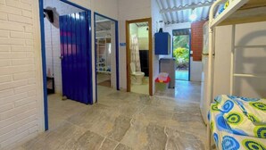 A room with a tiled floor, a bed with a patterned bedspread, a fan, and a door leading to another room.