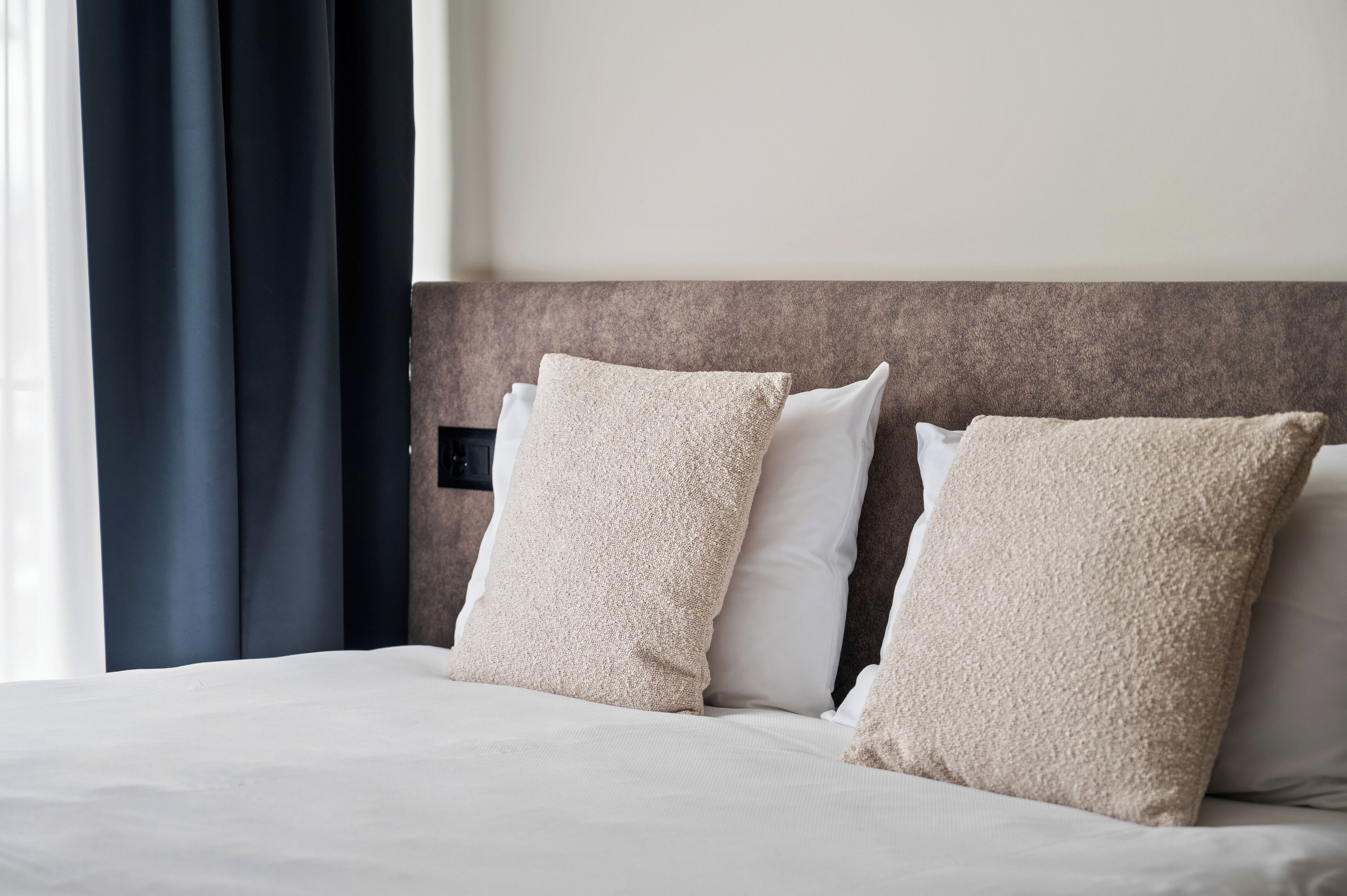 Una habitación de hotel con una cama, dos almohadas color beige, cabecera y ventana con cortinas.