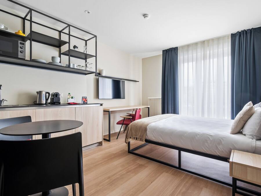 Habitación de hotel compacta con cama, escritorio, silla, una mesita y una zona de kitchenette.