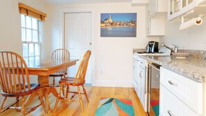 A kitchen with a wooden dining table, chairs, white cabinets, and a painting of a coastal town.