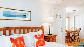 A neatly made bed with white bedding and orange pillows, a wooden headboard, a bedside table with a lamp, a framed picture on the wall, and a dining area with chairs and a table.