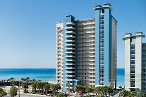 A high-rise hotel with multiple balconies, located near a beach with palm trees and a clear blue sky. - Grandview East 1002 (Panama City Beach)
