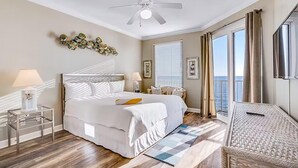 A hotel room with a bed, a ceiling fan, a window with blinds, a balcony with curtains, and a TV on the wall. - Grandview East 1002 (Panama City Beach)