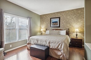 A bedroom with a bed, two bedside tables with lamps, a large window with curtains, a wall-mounted picture, and a wooden bench.