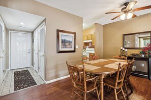 A dining area with a wooden table and chairs, a ceiling fan, and a kitchen in the background.