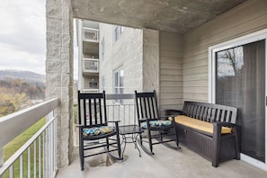 A balcony with two rocking chairs and a bench, a table, and a view of a building and landscape. - Free Dollywood Tkt | Mtn View Condo | Pigeon Forge | Sleeps 6 | Indoor Pool (Pigeon Forge)