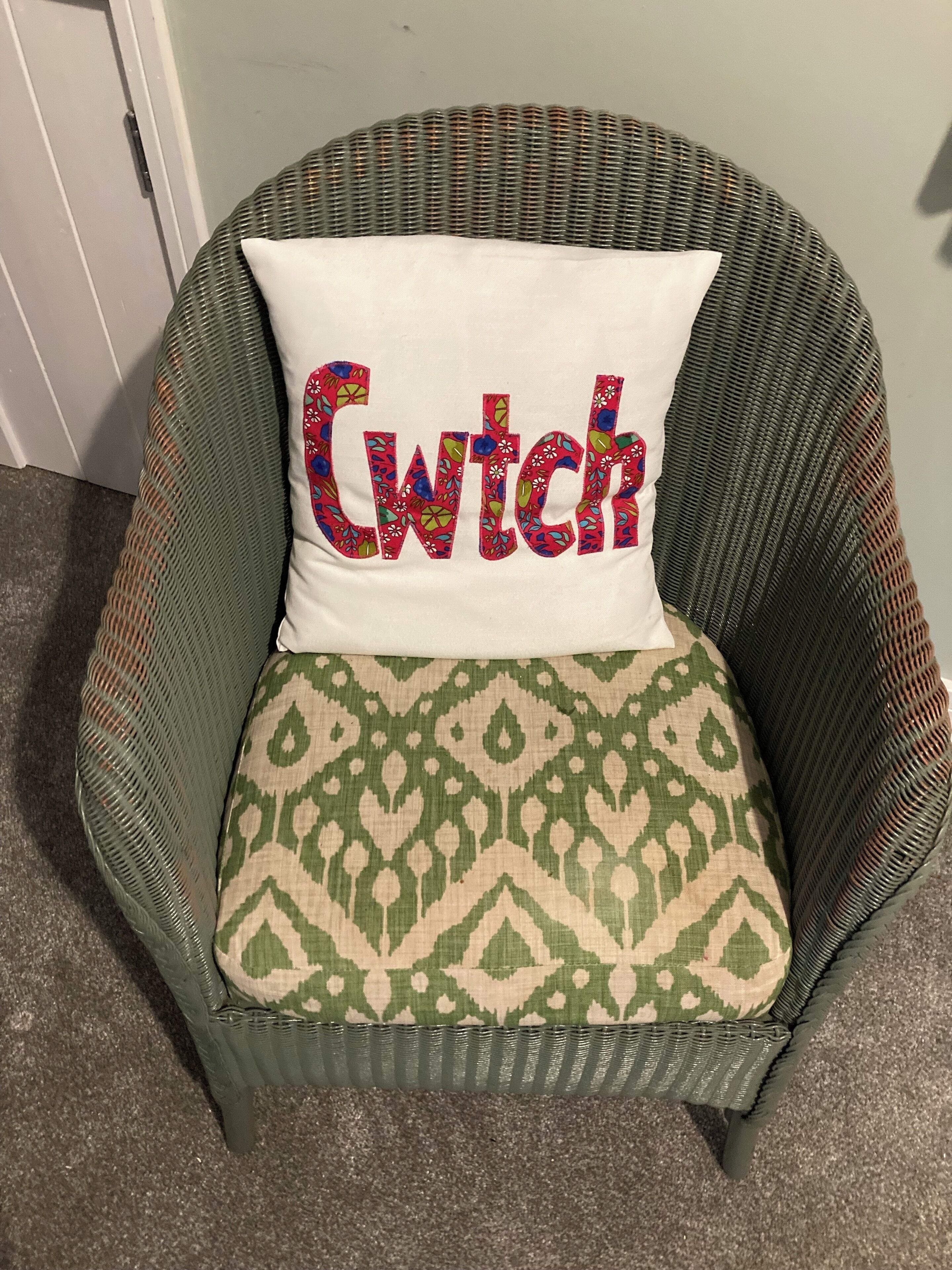 A wicker chair with a cushion that has a geometric pattern and a pillow with the word "Cwitch" embroidered in colorful letters.