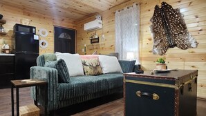 Interior acogedor de una cabaña con techo de madera, un sofá verde con cojines, un refrigerador negro y una maleta antigua.
