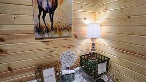 Interior de una cabaña de madera, con pared de madera, un cuadro de un ciervo, una mesa con un marco de fotos, una lámpara, un adorno y una cesta verde.