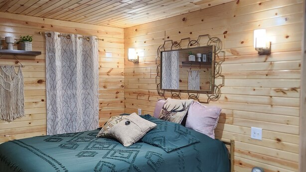 Habitación con paneles de madera, una cama, una mesita de noche, un espejo adosado a la pared y un estante pequeño con objetos decorativos.